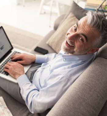Cheerful confident businessman at home sitting on the couch and working with a laptop, he is smiling at camera