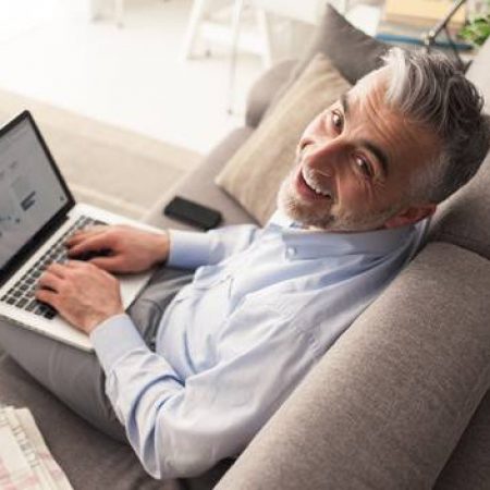 Cheerful confident businessman at home sitting on the couch and working with a laptop, he is smiling at camera